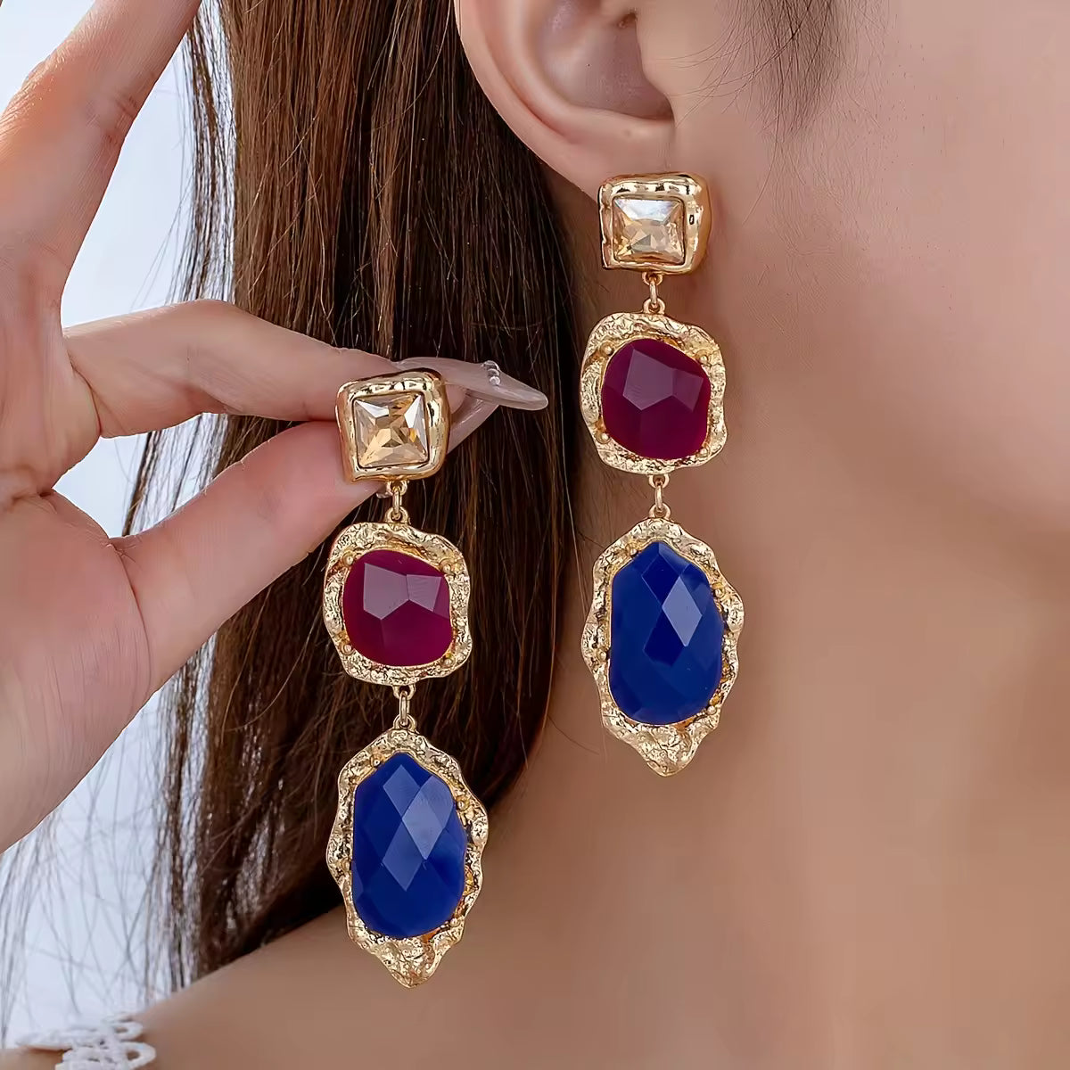 Gold earrings with red and blue gemstones worn by a person.
