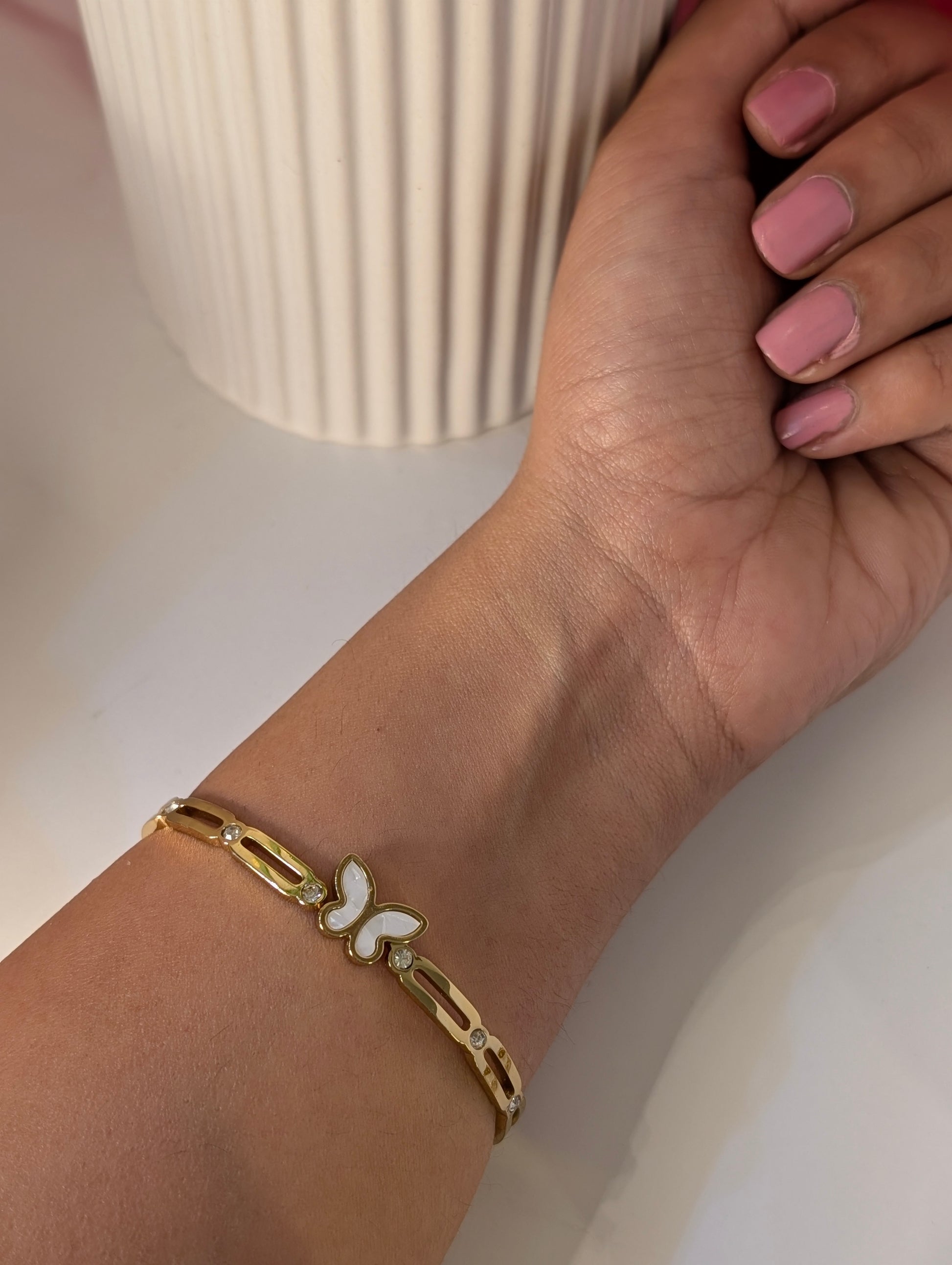 Gold bracelet with a butterfly charm on a wrist, against a neutral background