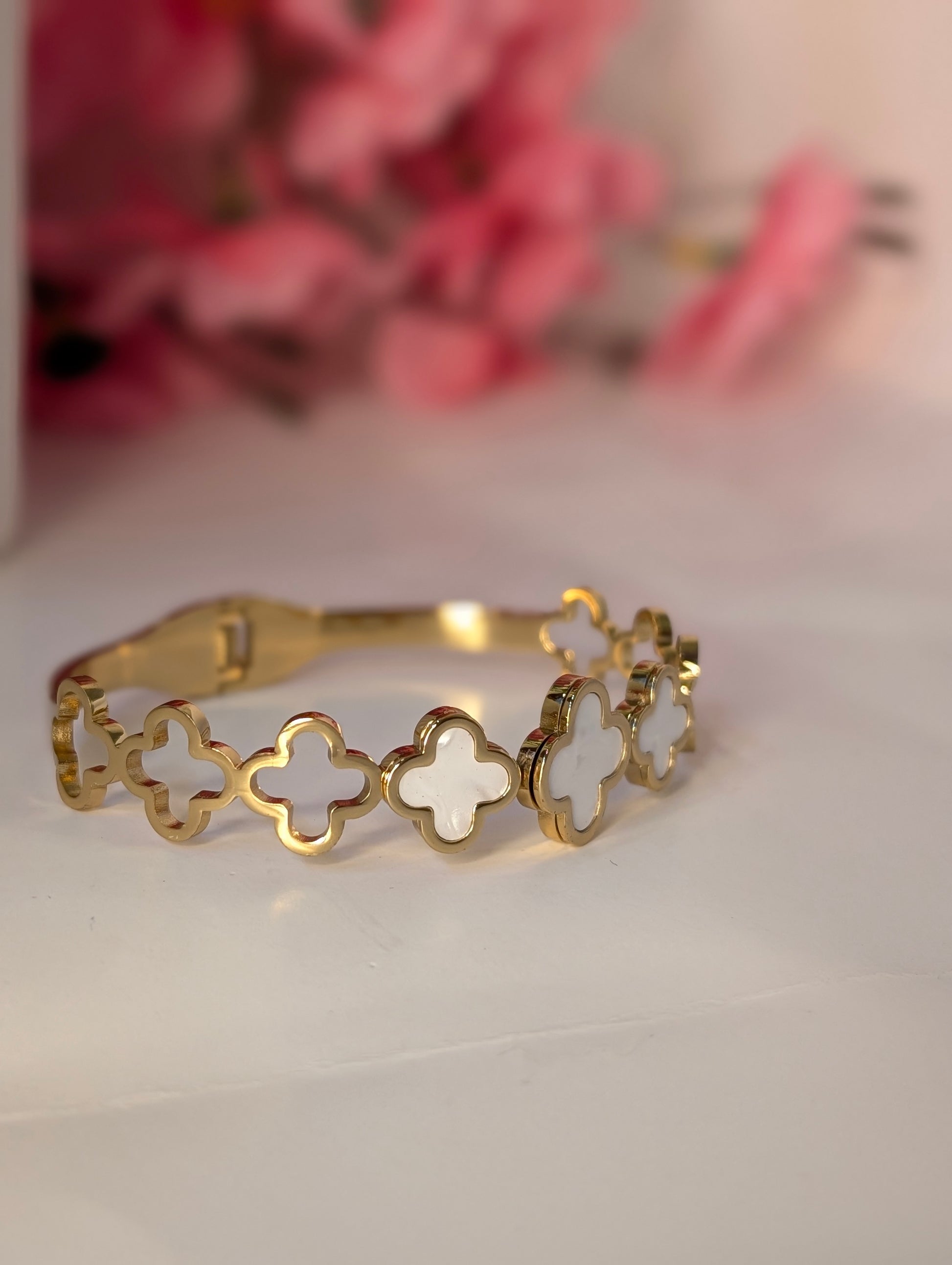Gold bracelet with clover designs on a white surface with pink flowers in the background